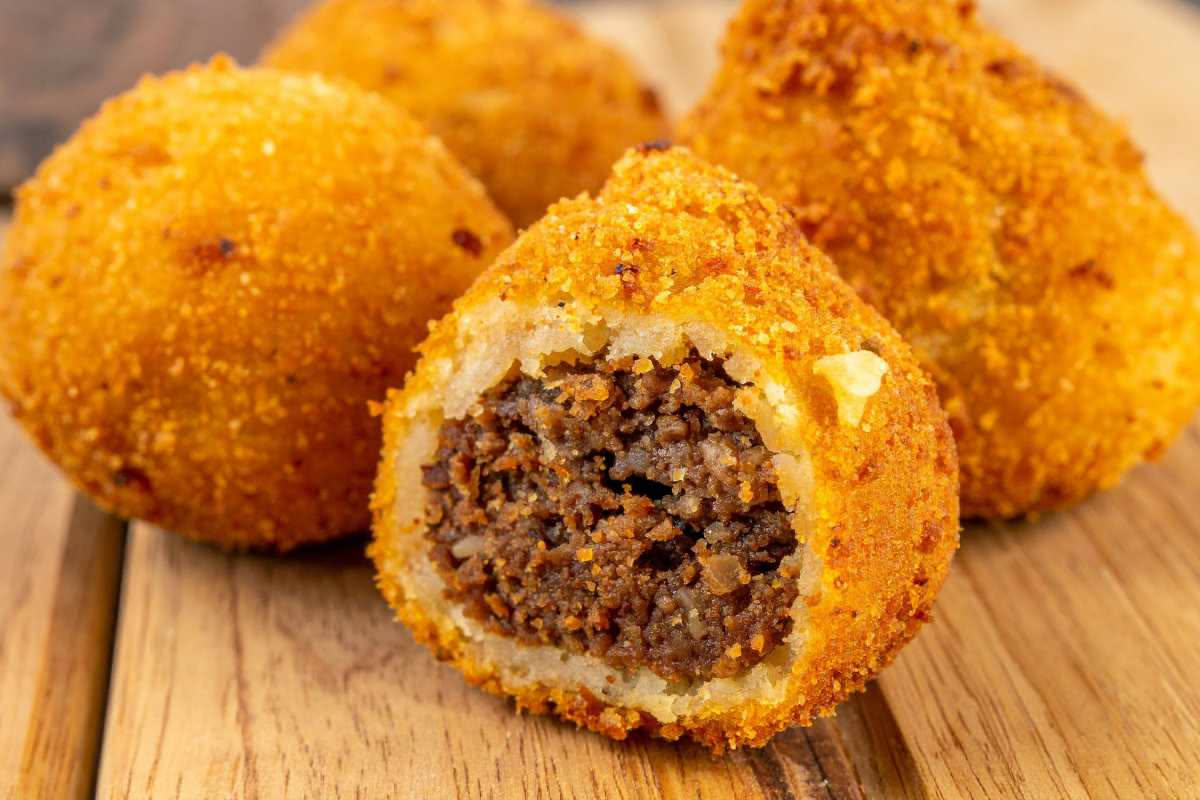 Tre arancini dorati e croccanti su un piatto bianco, tagliati a metà per mostrare il ripieno giallo di riso, ragù rosso e piselli verdi, con una salsa di pomodoro accanto