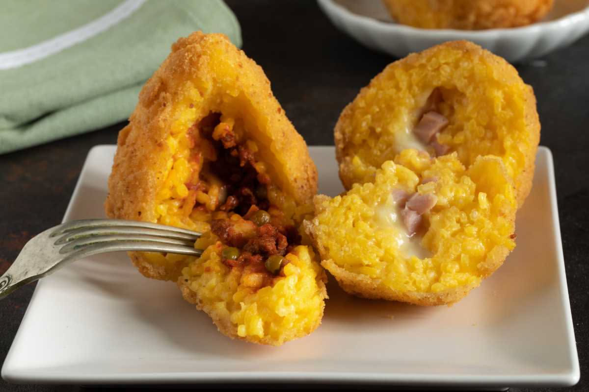 Tre arancini dorati e croccanti appena fritti, poggiati su carta bianca. All interno si vede il ragù marrone e la mozzarella filante. Accanto una ciotola di salsa al pomodoro.