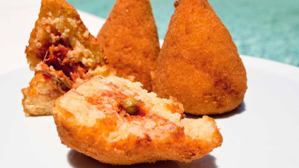 Tre arancini dorati e croccanti tagliati a metà, riempiti di ragù scuro e formaggio filante, su un piatto bianco con una salsa rossa accanto