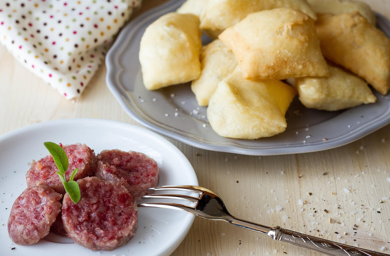 Tre arancini ragusani di forma ovale allungata, fritti e dorati, disposti su un piatto bianco con una ciotola di ragù di carne sullo sfondo
