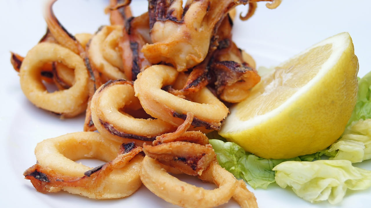 Calamaretti dorati e croccanti appena fritti in uno scolapasta, con limone fresco tagliato e sale grosso su piatto bianco