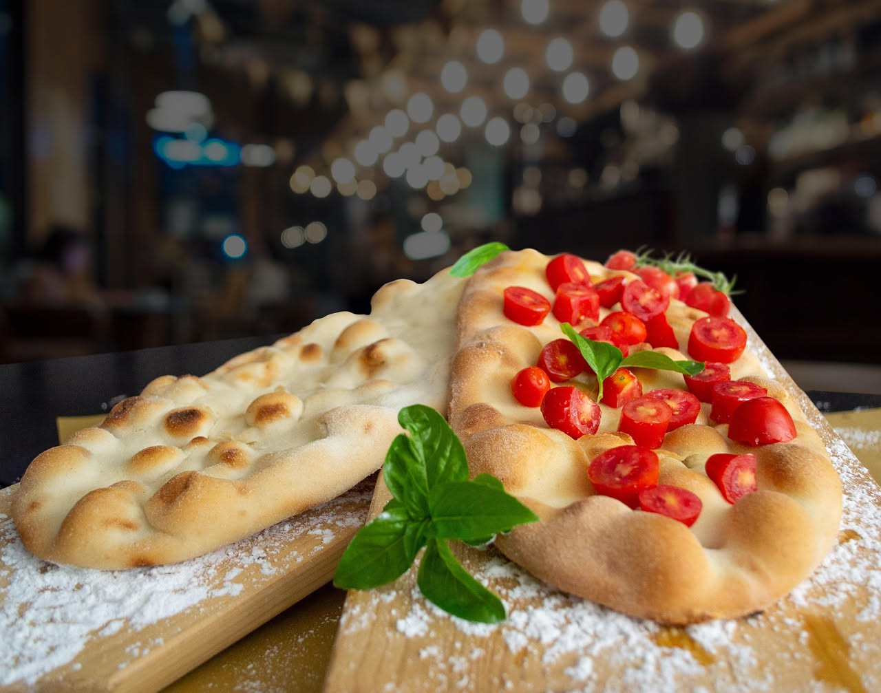 Focaccia napoletana appena sfornata, dorata e lucida, con pomodori rossi e origano verde sparsi sulla superficie, tagliata in rettangoli, su una tavola di legno bianco