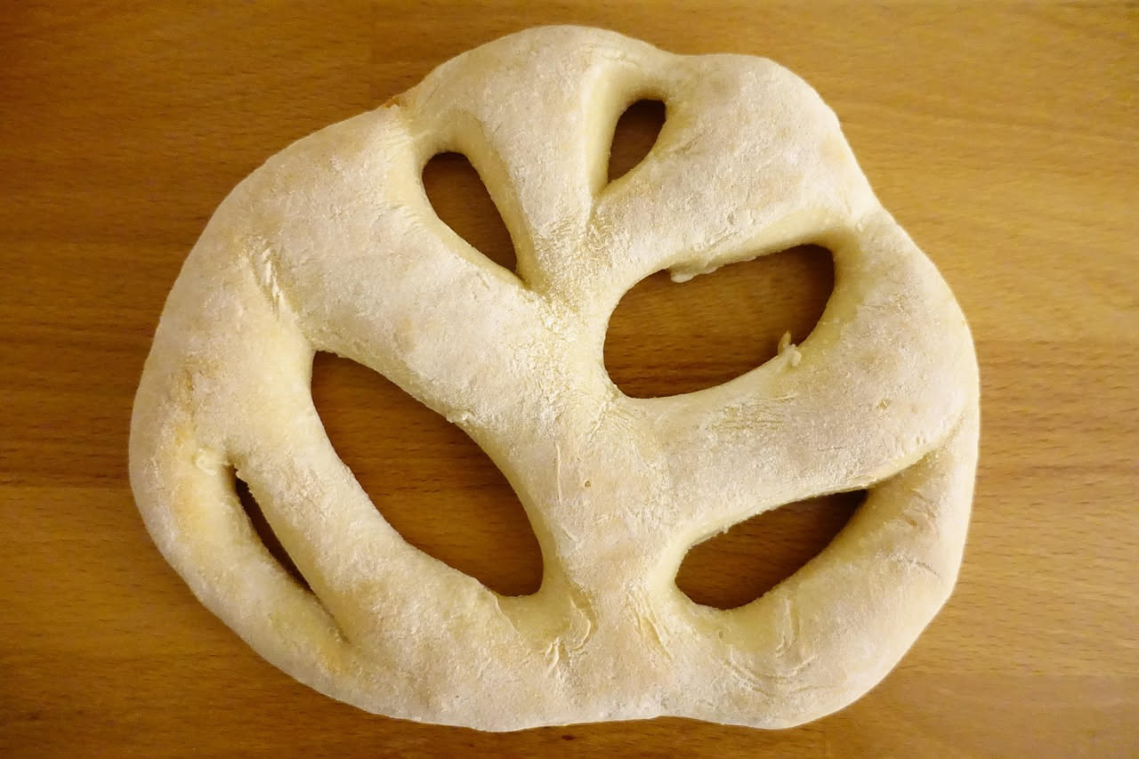 Focaccia riccia appena sfornata con superficie ondulata dorata, olio brillante nelle pieghe, rosmarino fresco sparso in superficie su teglia di carta da forno