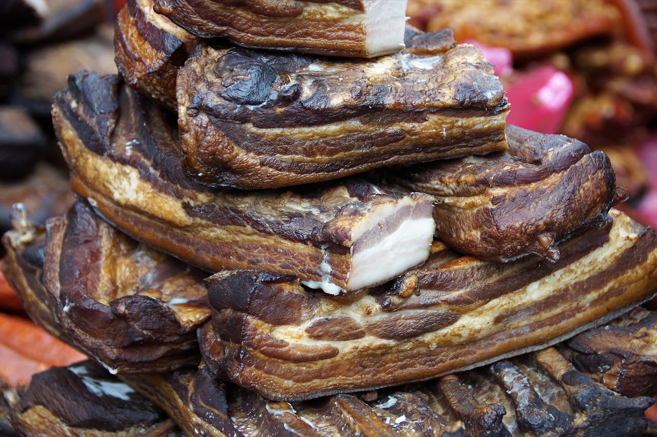 Fette di lardo affumicato di colore ambrato con striature marmorizzate, su tavolo di legno scuro con rametto di rosmarino e chicchi di pepe nero accanto