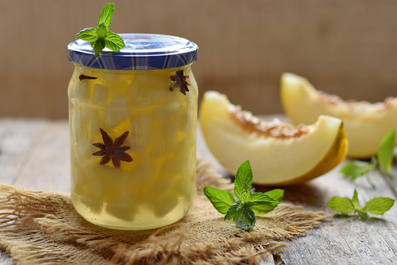 Bottiglia di vetro con liquore trasparente color ambra contenente fettine di melone, posta su una superficie di legno con melone fresco tagliato accanto e vista del sole al tramonto