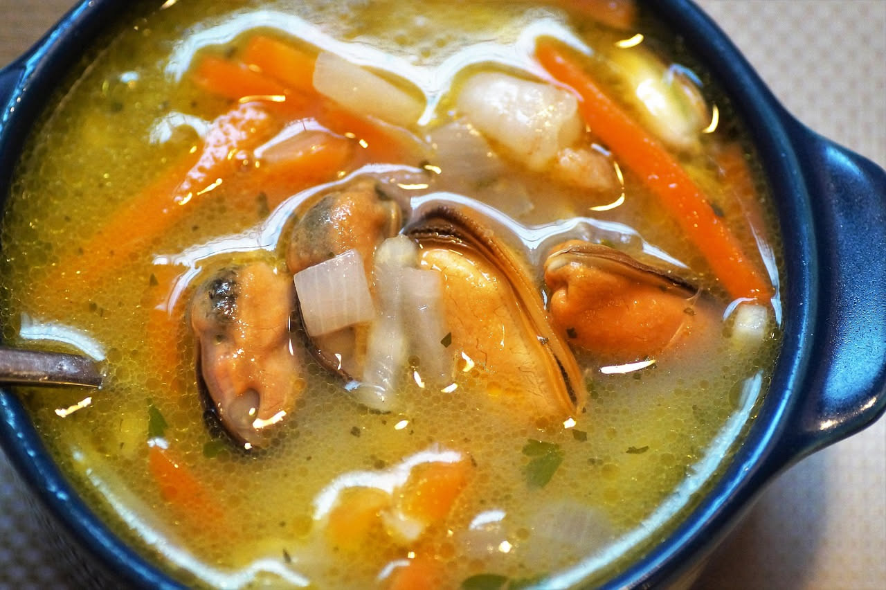 Minestra marina in ciotola bianca con cozze nere, vongole, brodo trasparente, carota e sedano visibili, cucchiaio di legno sul bordo