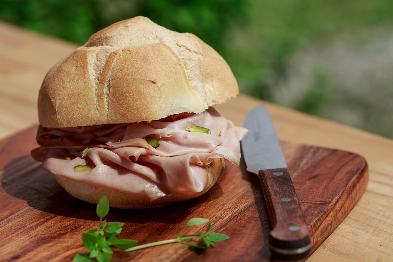 Fette di mortadella di Palermo rosa e grigia su un tagliere di legno con pepe nero grosso e rametti di rosmarino accanto