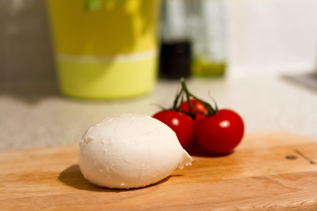 Mozzarella di bufala bianca, liscia e lucida in una ciotola di ceramica con acqua di conservazione, accanto a pomodori rossi e foglie di basilico fresco