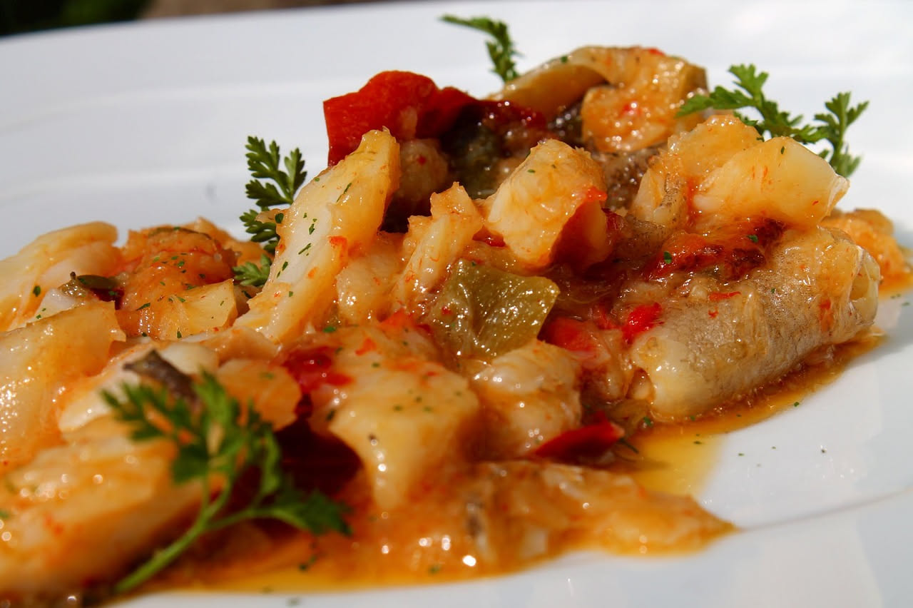 Filetti di nasello in carpione su un piatto bianco, ricoperti di verdure sottili e liquido agrodolce, con cipolla bianca e rosmarino fresco
