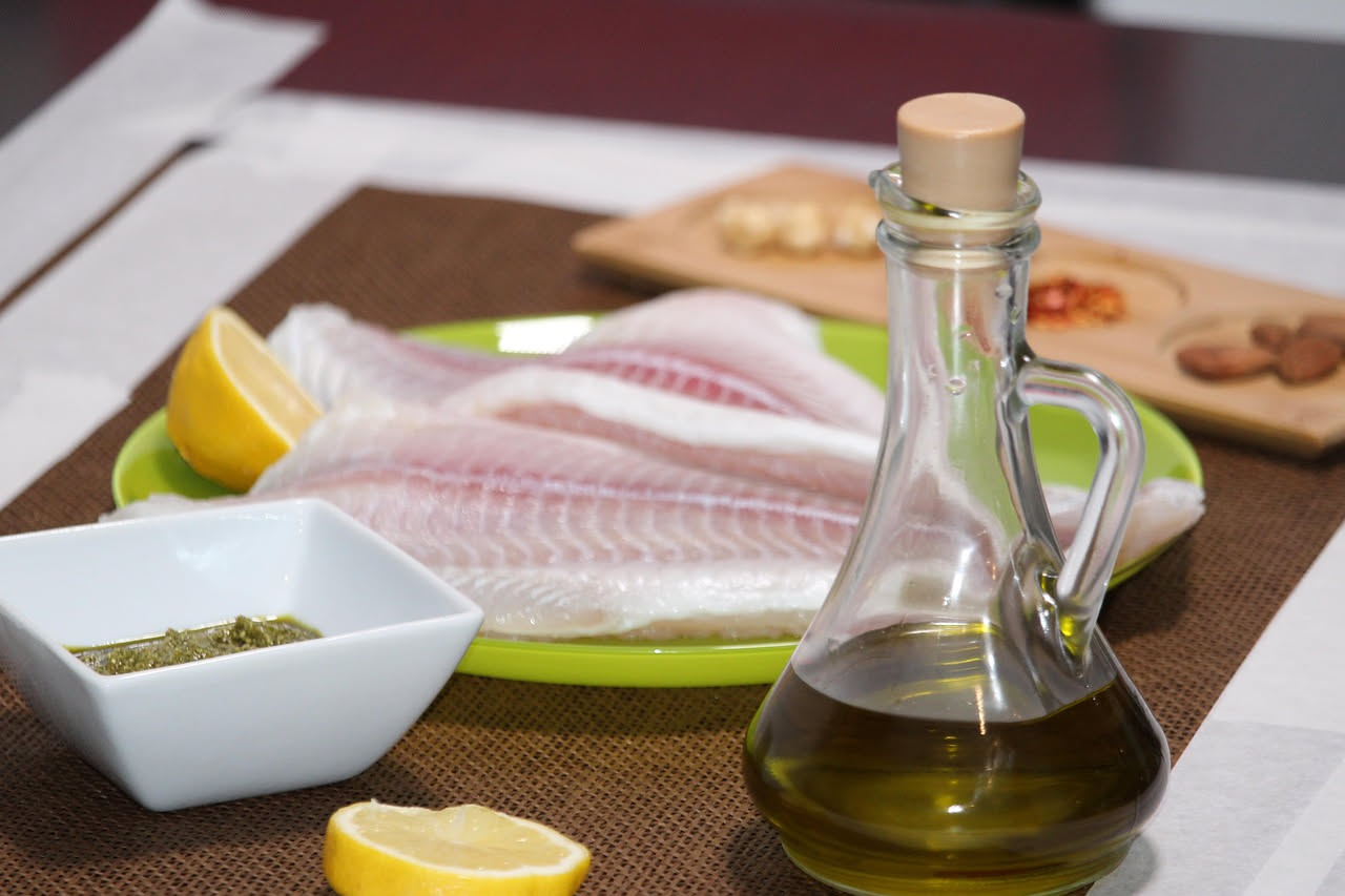 Nasello fresco filettato su piatto bianco con fette di limone giallo, olio d'oliva gocce trasparenti, peperoncino rosso e prezzemolo verde sfumato