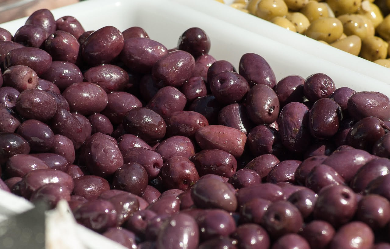 Olive nere di Gaeta in barattolo di vetro con salamoia, acqua, sale e foglie di alloro visibili, disposte su un tavolo di legno chiaro con torchon bianco
