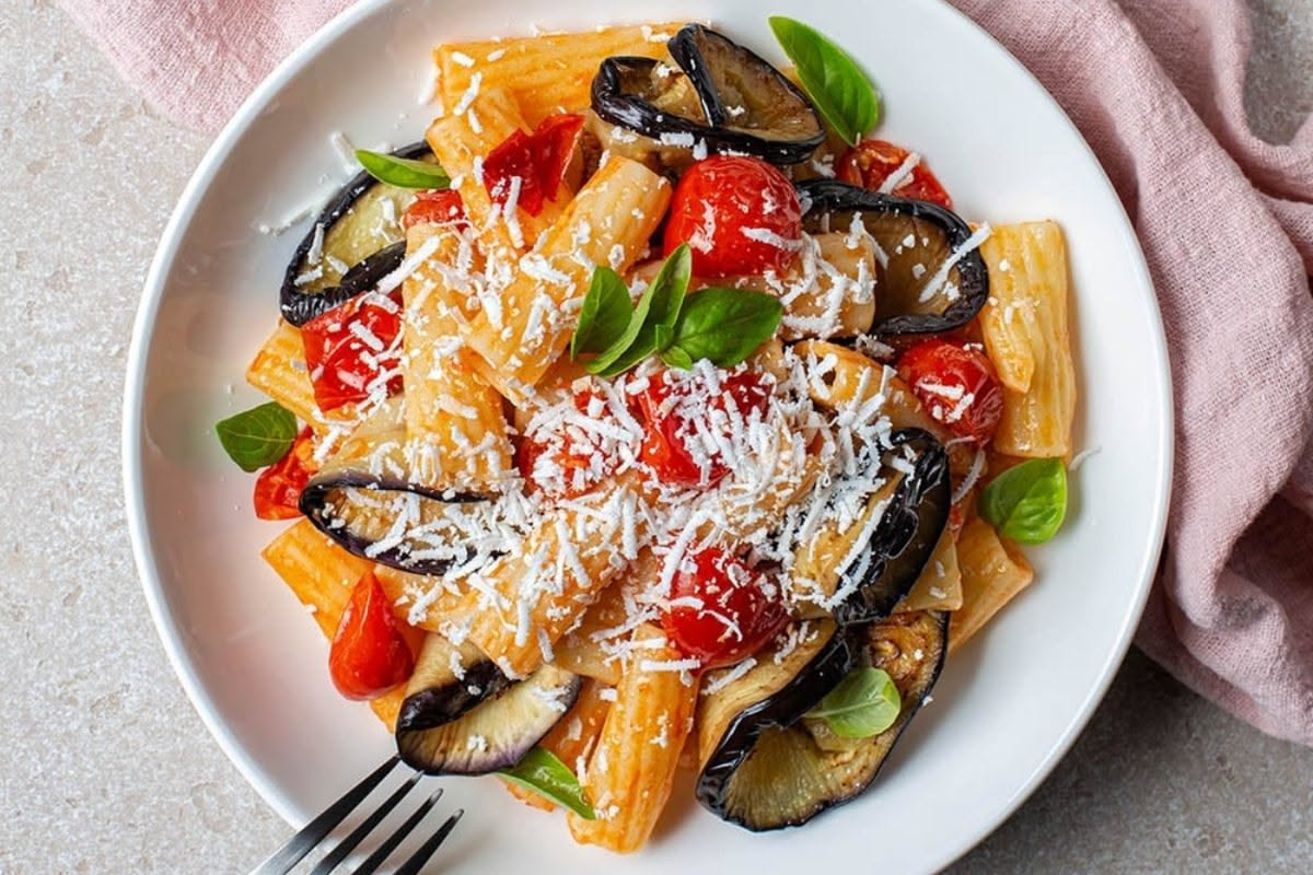 Piatto di pasta alla Norma con melanzane dorate, sugo rosso di pomodoro, ricotta salata bianca grattugiata e foglie di basilico fresco