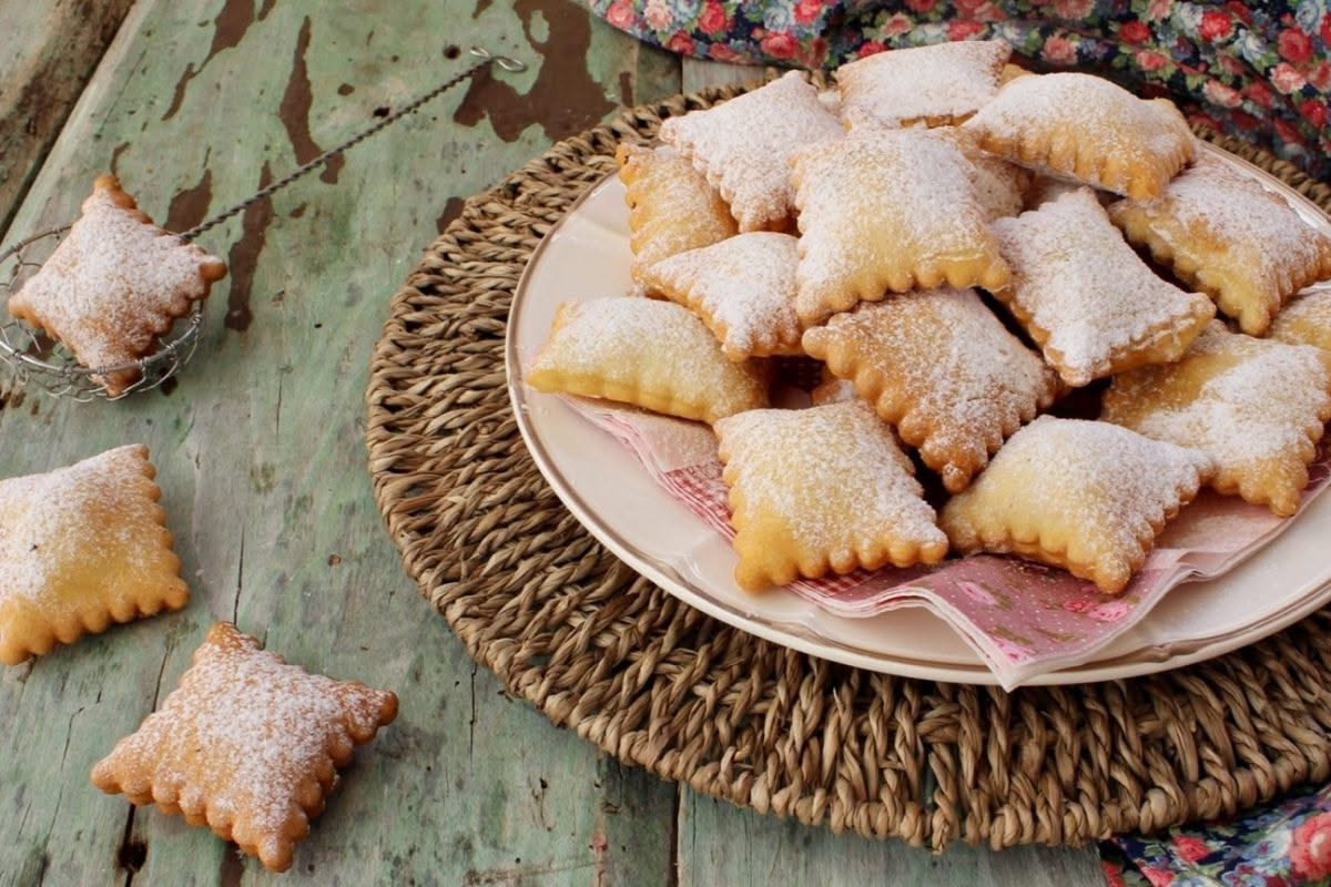 Pasta fritta cu' la ricotta appena fritta su piatto bianco, dorata e croccante, cosparsa di zucchero fine, con una coppetta di ricotta fresca accanto