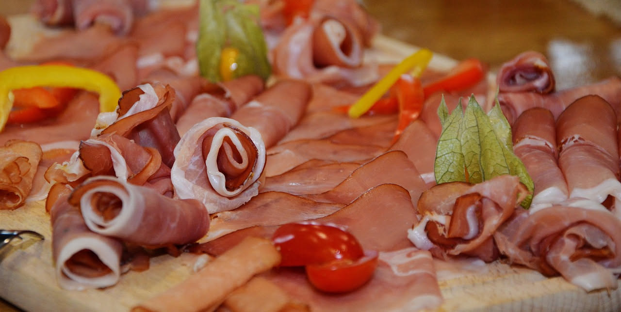Tagliere in legno rettangolare con affettati misti disposti a ventaglio: prosciutto crudo rosa, mortadella chiara, speck scuro, soppressata rosso intenso, culatello beige, su fondo bianco con pane tos
