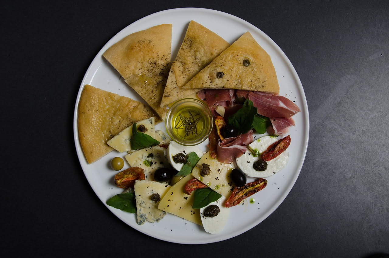 Tagliere di legno con affettati misti disposti in spirale: prosciutto crudo rosato, speck affumicato color mogano, pancetta striata, guanciale, salame rosso scuro. Contorno di pane tostato e olive ver