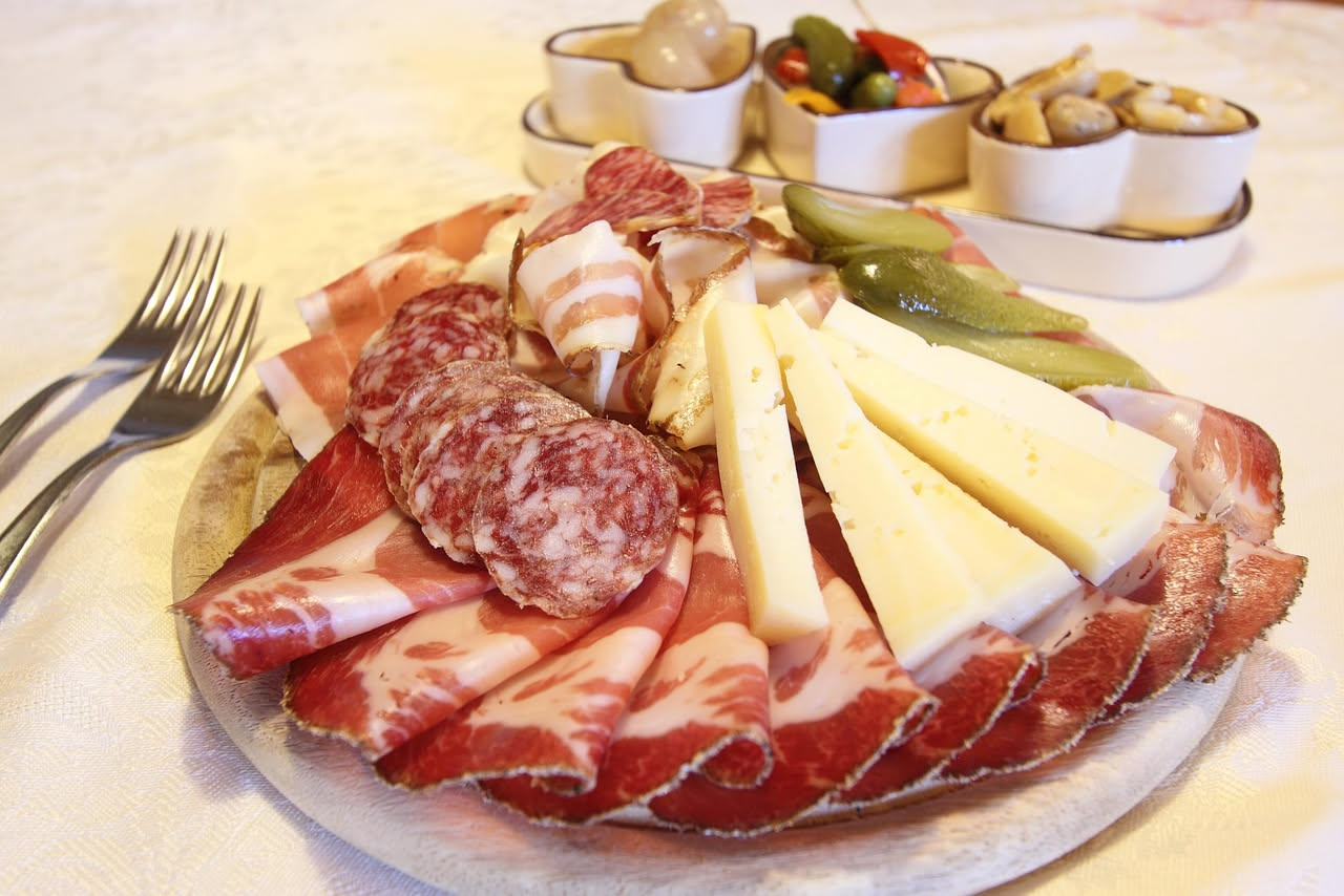 Affettati misti su un tagliere di legno chiaro: fette di prosciutto rosa tenue, salame rosso scuro, mortadella con grani di pepe visibili, coppa marmorizzata. Accanto, pane toscano e un calice di vino