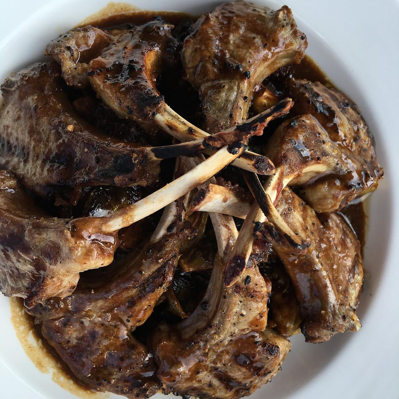Piatto fondo bianco con pezzi di agnello rosso scuro immersi in sugo marrone scuro, contornato da verdure morbide, cipolla bianca e carota arancione, vapore visibile, cucchiaio d'argento appoggiato su