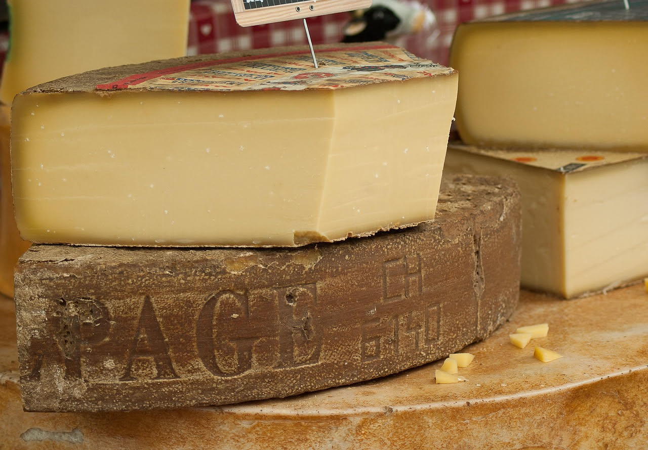 Fette di Asiago d'allevo color paglierino disposte a ventaglio su piatto bianco, con cristalli visibili sulla superficie e una lama di coltello in acciaio sullo sfondo