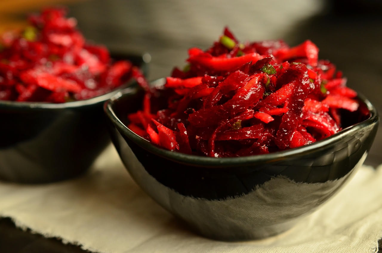 Barbabietola affettata in agrodolce in una ciotola di vetro, rosso intenso, con fette sottili coperte da sciroppo trasparente con riflessi dorati, guarnita con un rametto di prezzemolo fresco