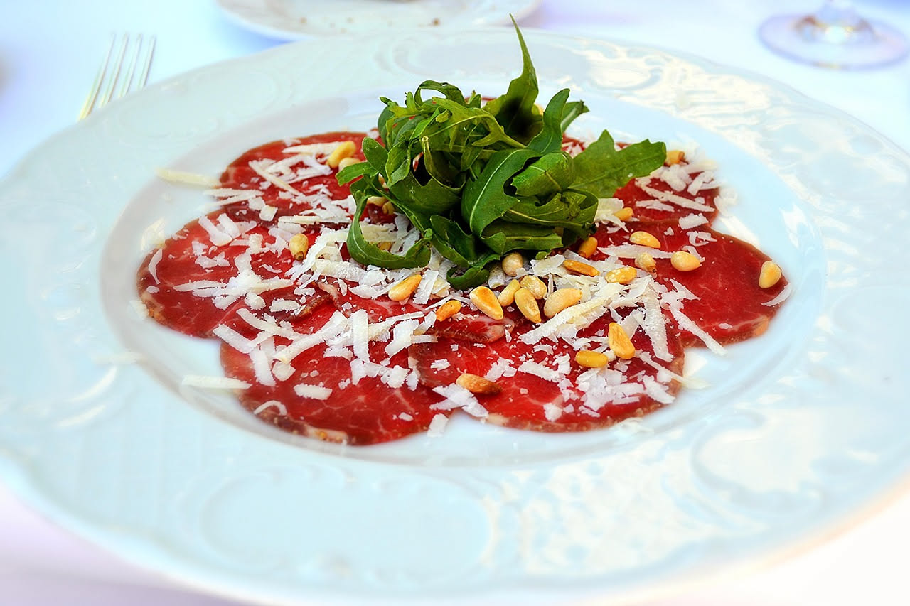 Fette sottili di bresaola di colore rosso scuro piegate su un piatto bianco, cosparse di olio extravergine dorato e gocce di limone fresco, con un rametto di rosmarino