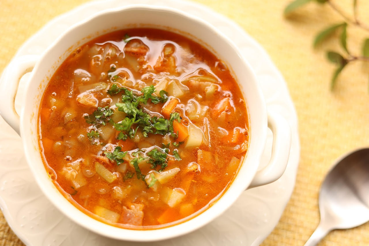 Brodetto pesarese in ciotola di ceramica bianca: brodo color ambra con pezzi di pesce, pomodoro e cipolla, accompagnato da fetta di pane tostato croccante bagnata nel brodo, tavolo di legno scuro