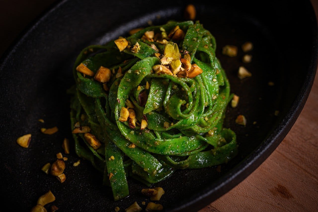 Piatto bianco con bucatini cremosi di colore verde chiaro dovuto al pistacchio, conditi in modo uniforme, con una spolverata di formaggio grattugiato bianco e qualche granello di pistacchio intero spa