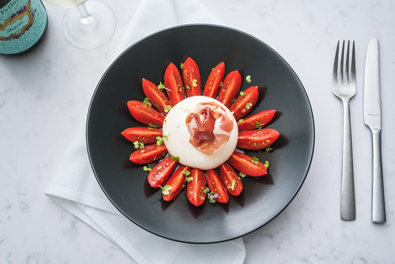 Piatto bianco con burrata cremosa al centro, pomodori rossi tagliati intorno, olio d'oliva dorato versato sopra, basilico fresco verde
