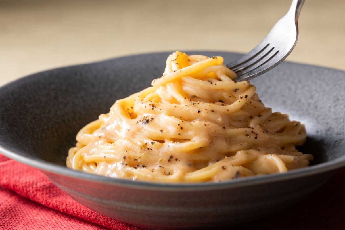 Un piatto bianco con una porzione di cavolfiore cremoso color giallo oro, ricoperto di pepe nero macinato grosso e scaglie di pecorino romano bianche; al centro, i florets si intravedono sotto la sals