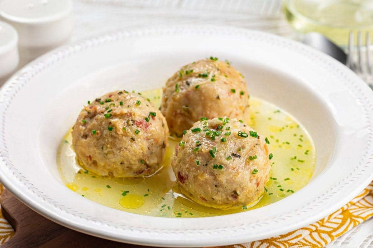 Canederli tondi e dorati in una ciotola di brodo trasparente freddo, con prezzemolo fresco e un filo di brodo ambrato visibile intorno agli gnocchi