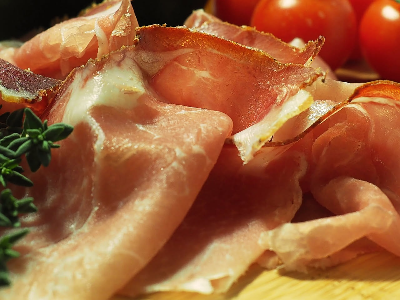 Affettato di capicola color rosso scuro su tagliere di legno con mozzarella fresca e pane carasau, accanto a bicchiere di vino rosso