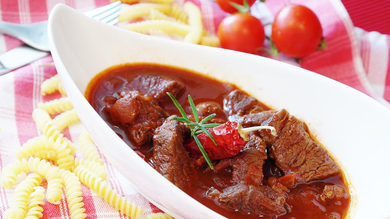 Piatto di capriolo in salmì: pezzi di carne scura e tenera immersi in sugo di vino rosso scuro, con carote e sedano visibili, servito in ciotola di ceramica bianca su tavolo di legno