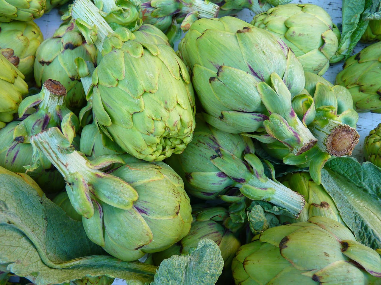 Carciofi interi verdi scuro disposti in una pentola di terracotta, con brodo trasparente, fette di aglio visibili e prezzemolo fresco tritato in superficie
