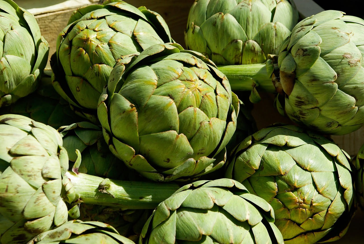 Carciofi interi cotti in pentola di terracotta, dorati e morbidi, con aglio e prezzemolo verde, serviti in una ciotola di ceramica rustica con un filo d'olio d'oliva lucente