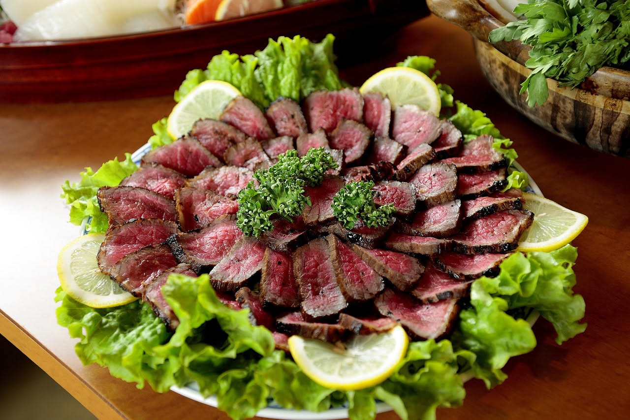 Piatto bianco con carpaccio di capriolo: fette sottili di carne color rosso intenso, disposte in cerchio, decorate con gocce di olio verde e scaglie di parmigiano, su sfondo neutro