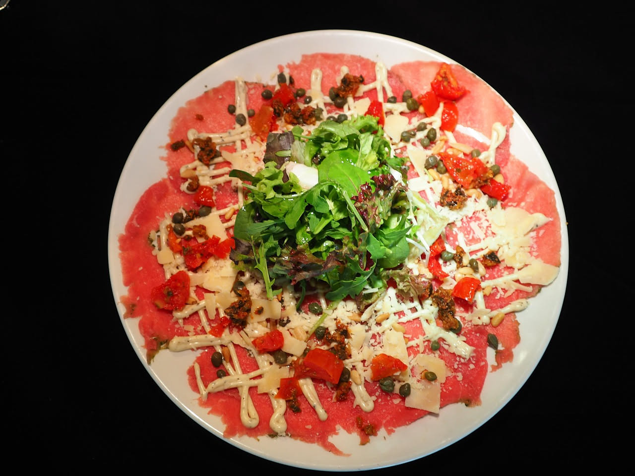 Piatto bianco con fette sottilissime e traslucide di manzo rosa pallido, coperte di olio extravergine lucido, scaglie di parmigiano e gocce di limone fresco, decorato con rucola verde