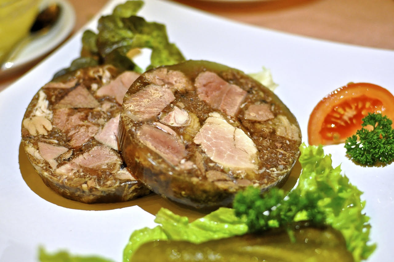 Fette di coppa di testa su un tagliere di legno, con gelatina trasparente intorno alla carne rosa, accanto a pane tostato e un coltello da cucina