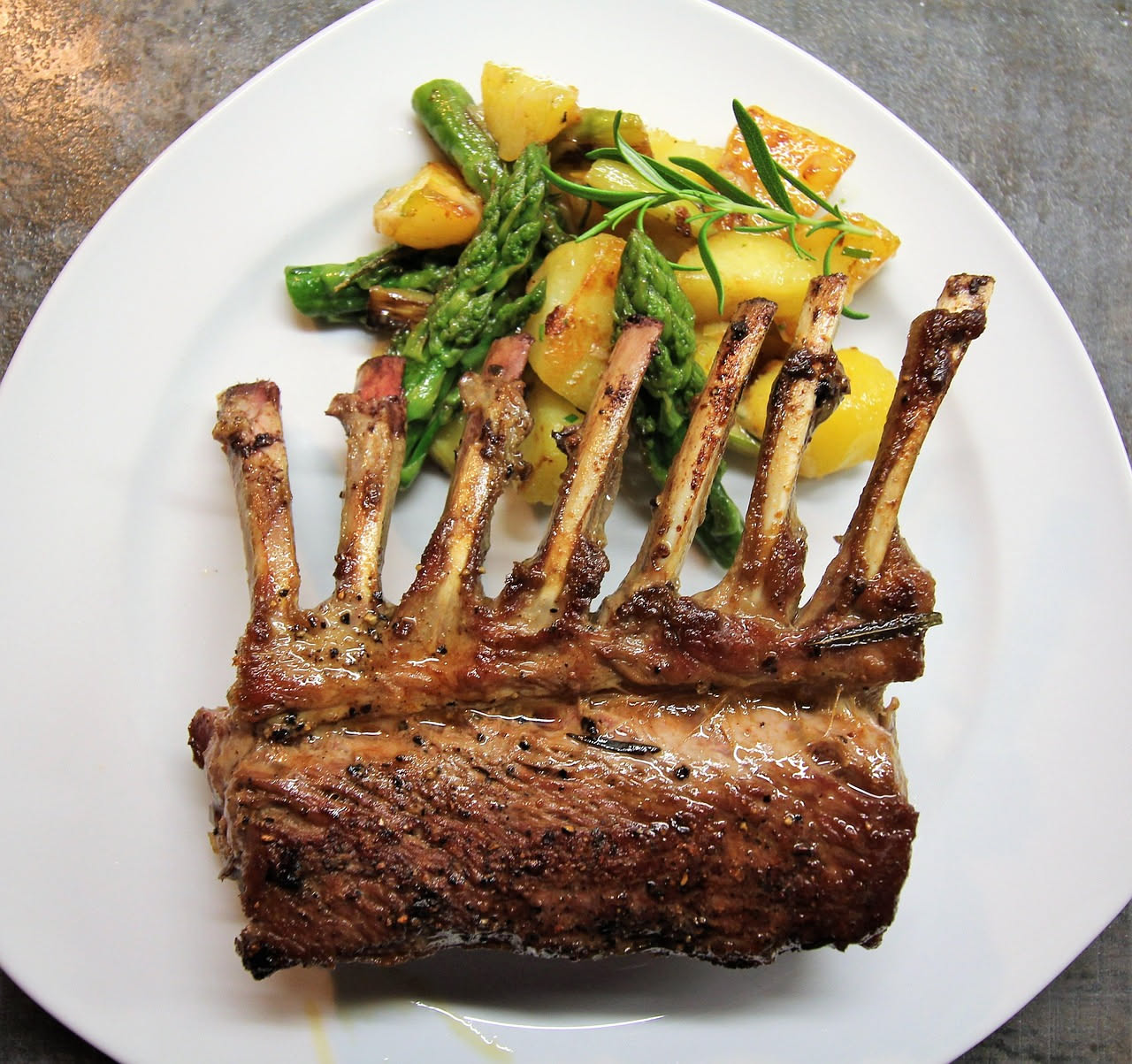 Quattro costolette di agnello dorate e rosate al centro, adagiate su un piatto bianco con contorno di limone fresco tagliato a spicchi e rametti di rosmarino