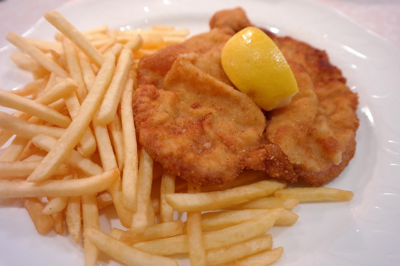 Cotoletta dorata e croccante su piatto bianco, accompagnata da una fetta di limone giallo e un pizzico di sale marino grosso sparso intorno