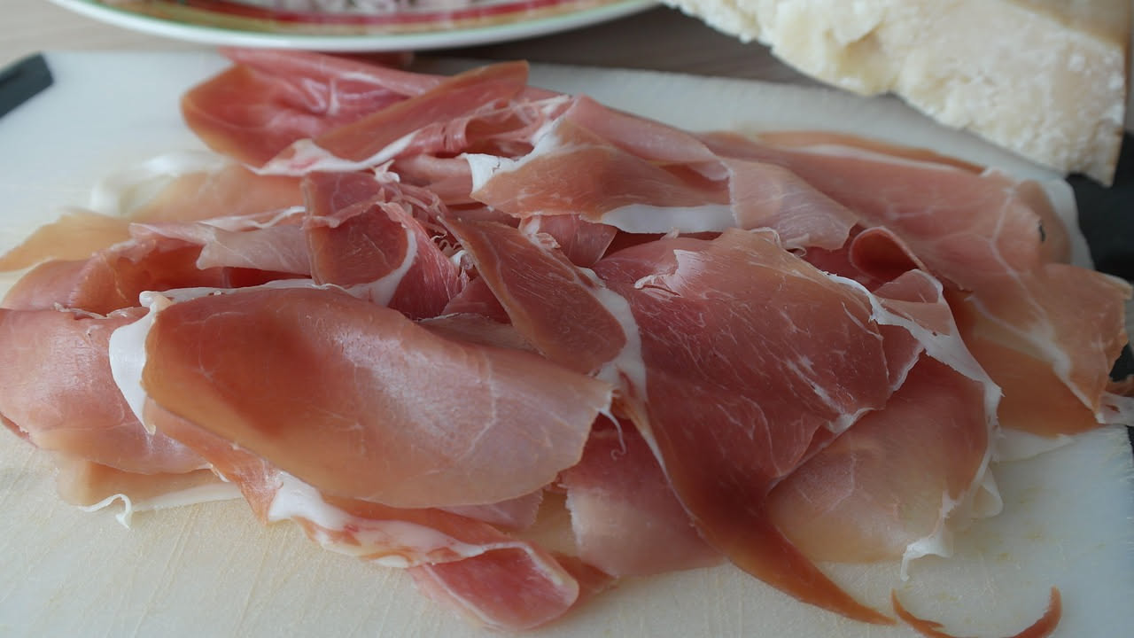 Fette sottili di culatello color rosso intenso disposte a ventaglio su piatto bianco con grissini e bicchiere di vino