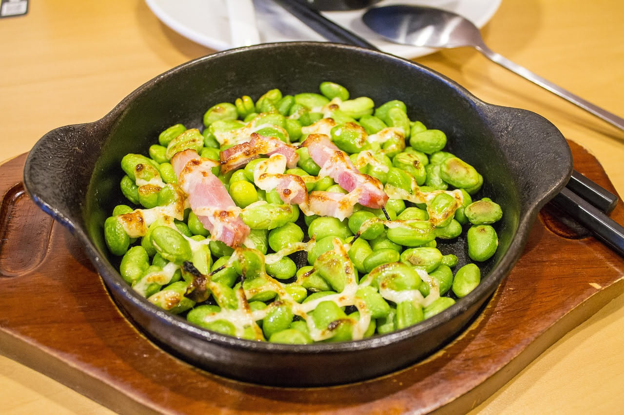 Piatto bianco con fave verdi appena sgusciate, croccanti e intere, alternate a scaglie sottili di formaggio giallo paglierino, guarnito con foglie di menta fresca e un pizzico di pepe nero.