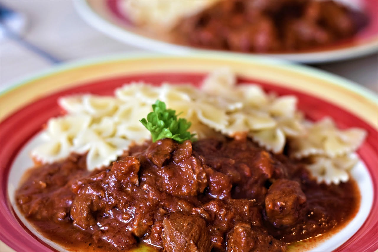 Fegatelli in umido in un piatto di ceramica beige, bagnati in salsa di pomodoro scuro, circondati da cipolla e erbe aromatiche, con pane tostato accanto