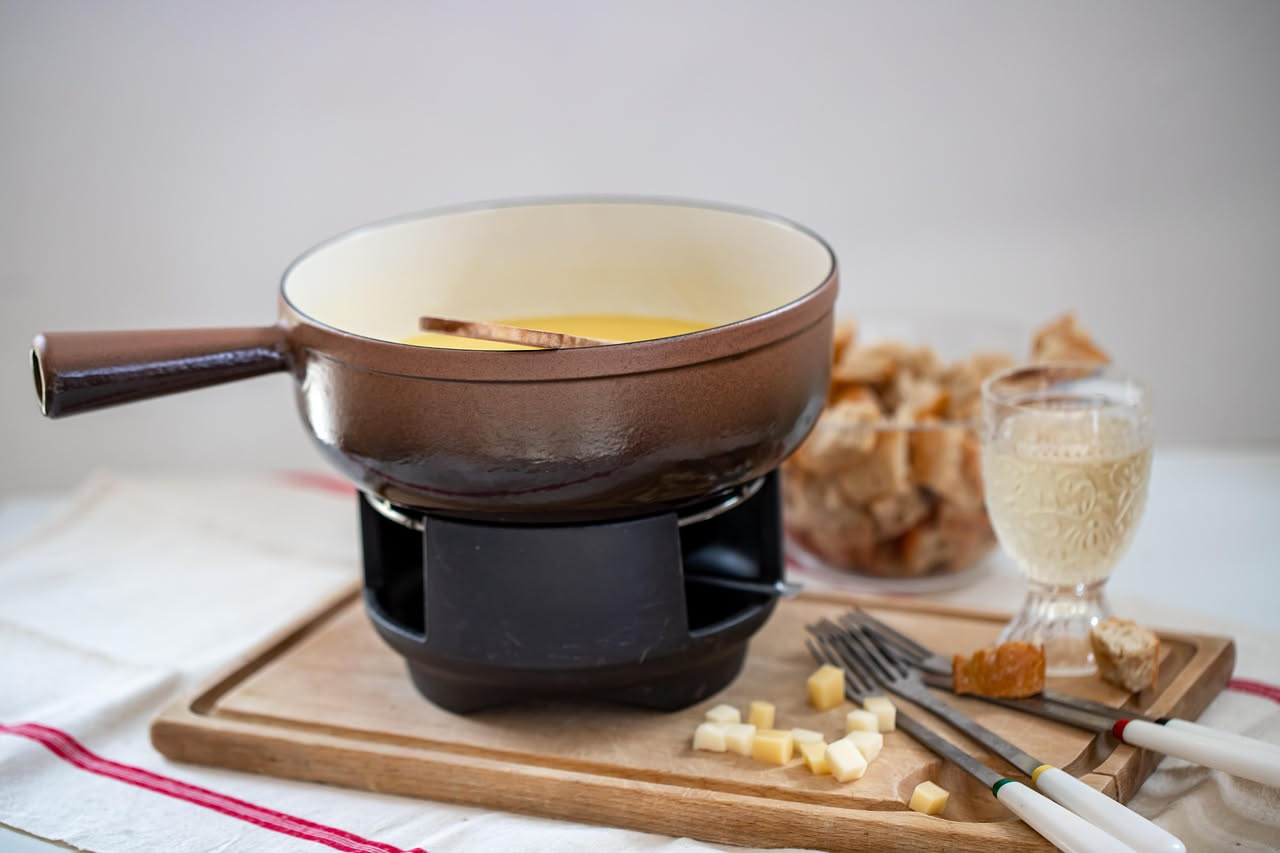 Fonduta calda e cremosa color giallo-beige in una pentola rotonda di ceramica bianca, bastoncini di pane tostato e fette di mela fresca disposte attorno su un tavolo di legno scuro