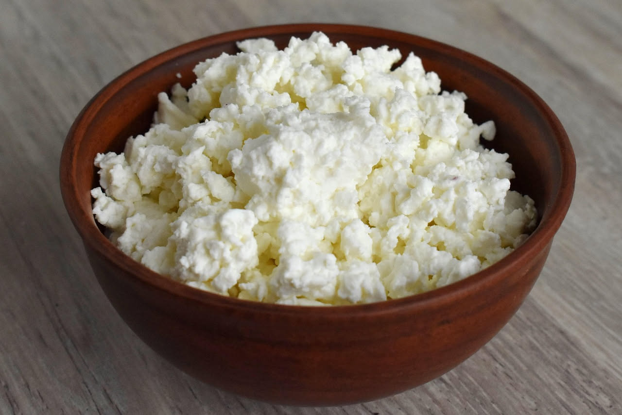 Ricotta fresca bianca e cremosa in una ciotola di ceramica, con accanto un cucchiaio di legno e una bottiglia d'aceto, su un tavolo di legno chiaro