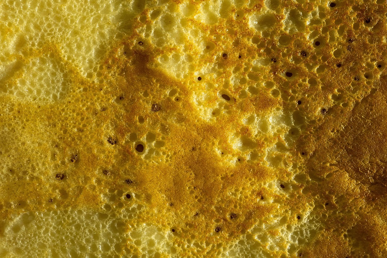 Frittelle di farina di mais dorate e croccanti impilate su un piatto bianco, ricoperte da uno strato sottile di zucchero, con una tazza di caffè sfocata sullo sfondo