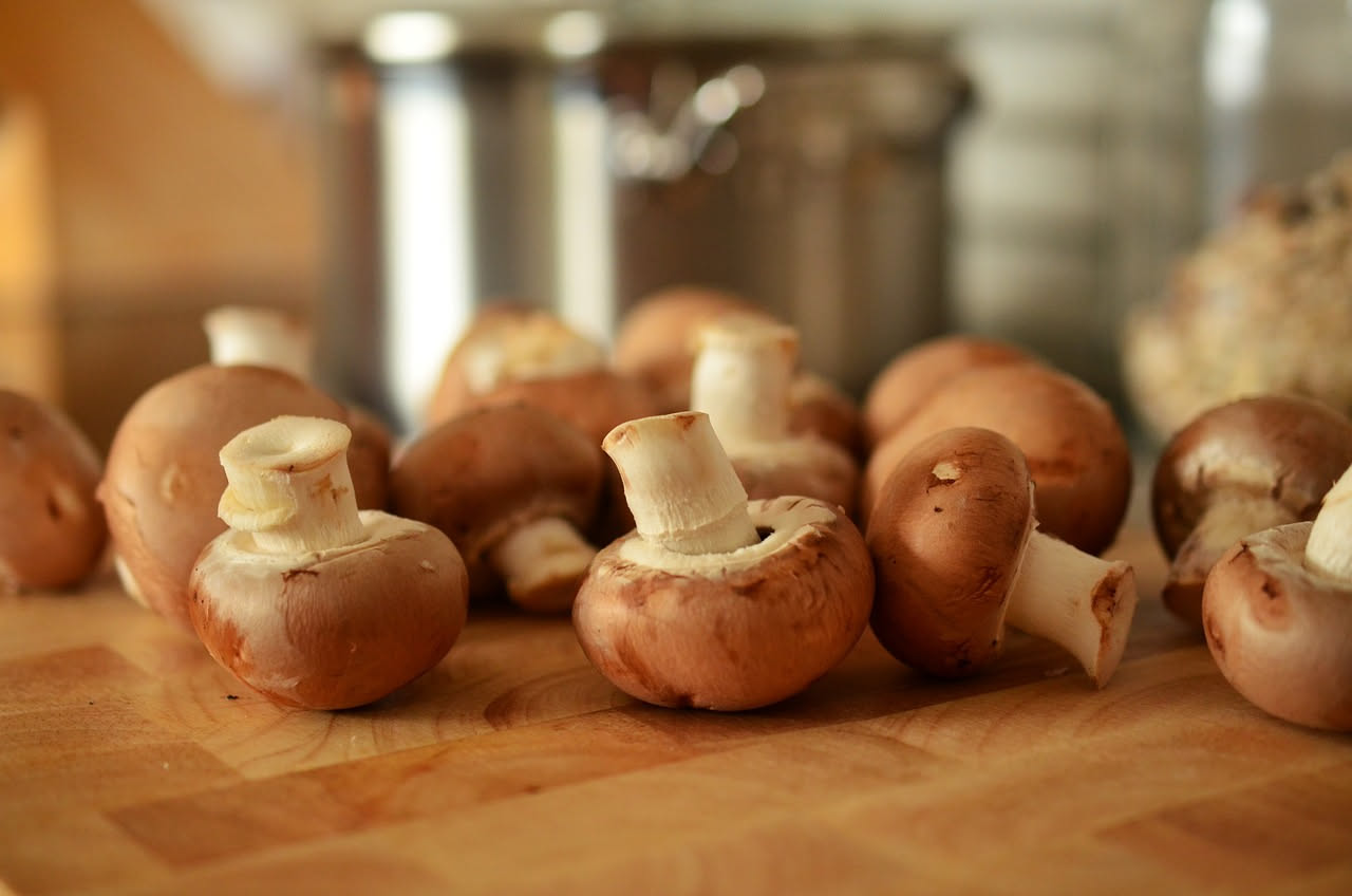 Vasetto di vetro trasparente con funghi bianchi e brunastri immersi in olio trasparente e rametti di timo, su tavolo di legno chiaro con spicchi d'aglio a lato