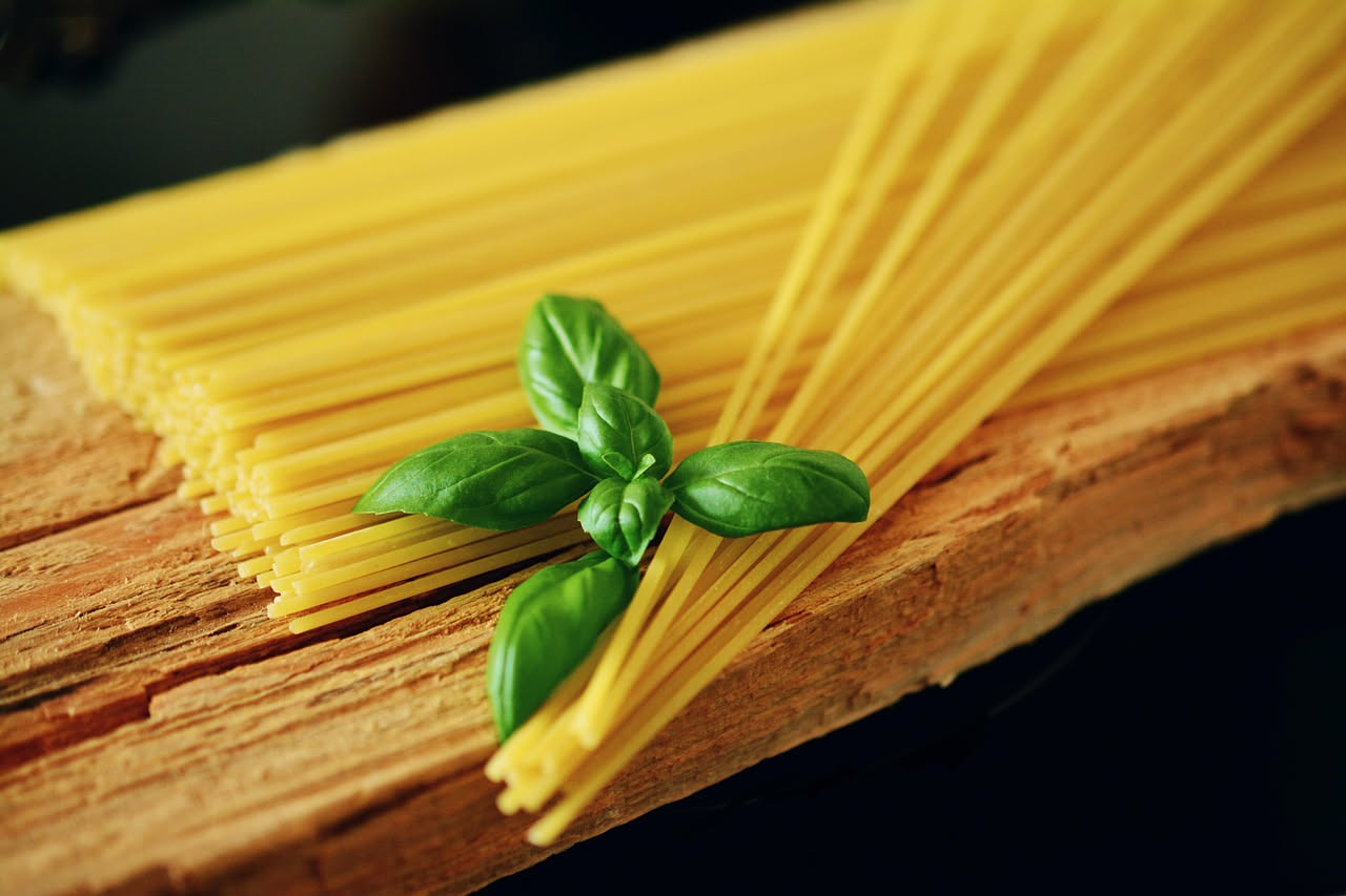 Un piatto bianco con fusilli ricoperti di foglie di basilico fresco verde brillante, olio dorato e aglio, con texture della pasta visibile, leggera spruzzata di parmigiano e un filo di olio a crudo