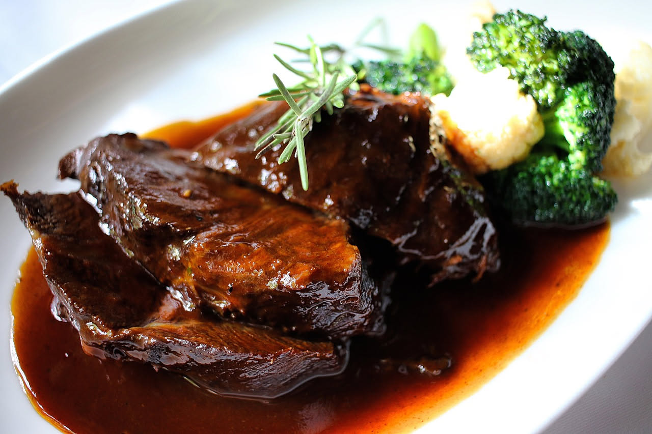 Guancia di manzo brasata intera, marrone scuro e lucida, su un piatto bianco con salsa di brasatura densa, contornata da carote e sedano cotti, con cucchiaio d'argento accanto e vapore tenue