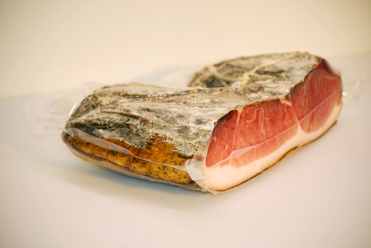 Fette sottili di guanciale senese disposte su un tagliere di legno, con la caratteristica colorazione rosata e i bordi di grasso bianco, accanto a pane toscano scuro e un calice di vino rosso