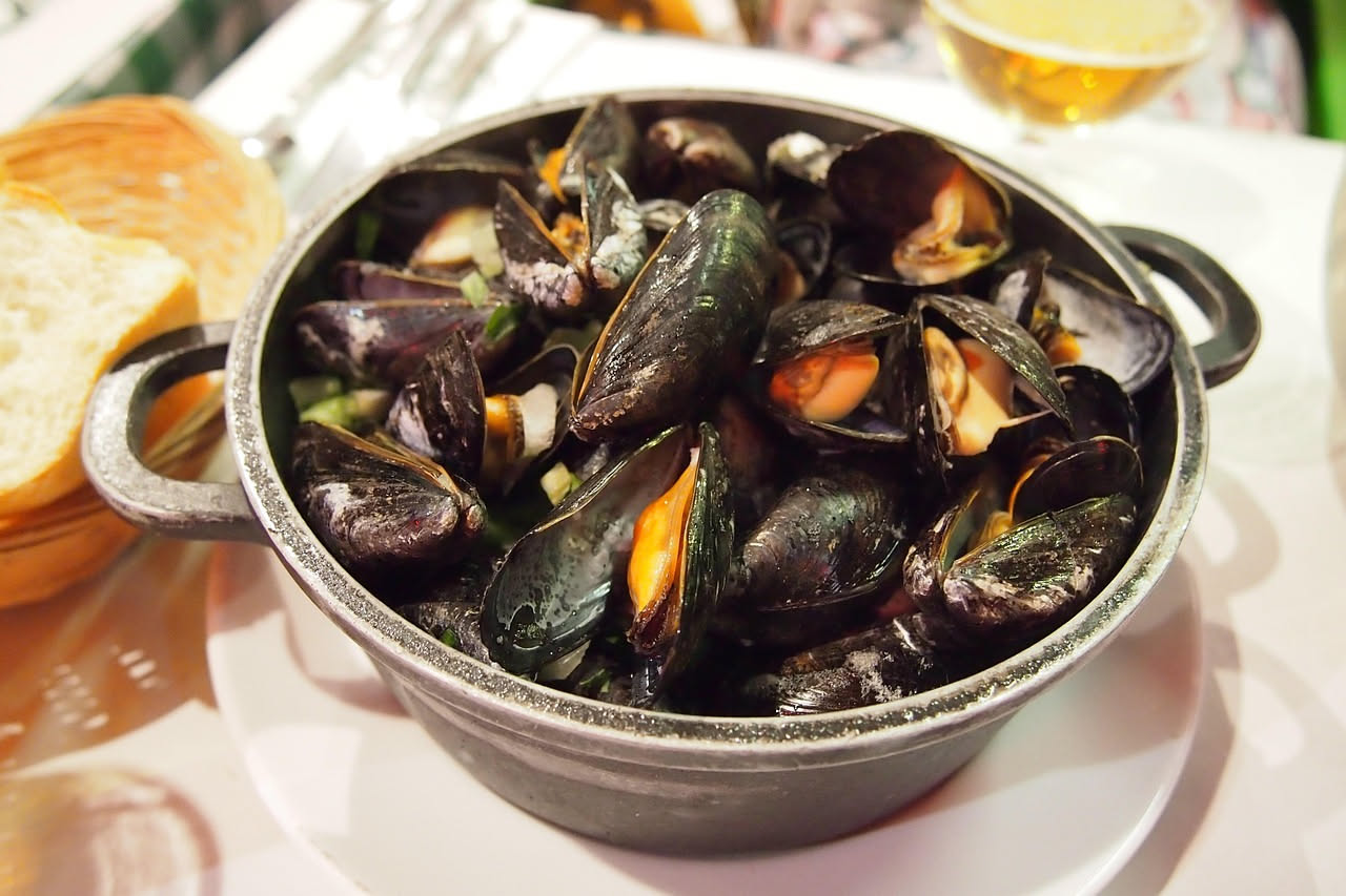 Ciotola di ceramica bianca con cozze aperte nere e guscio marrone scuro immerse in brodo trasparente con gocce di olio d'oliva e pepe nero visibile, pane tostato ai lati