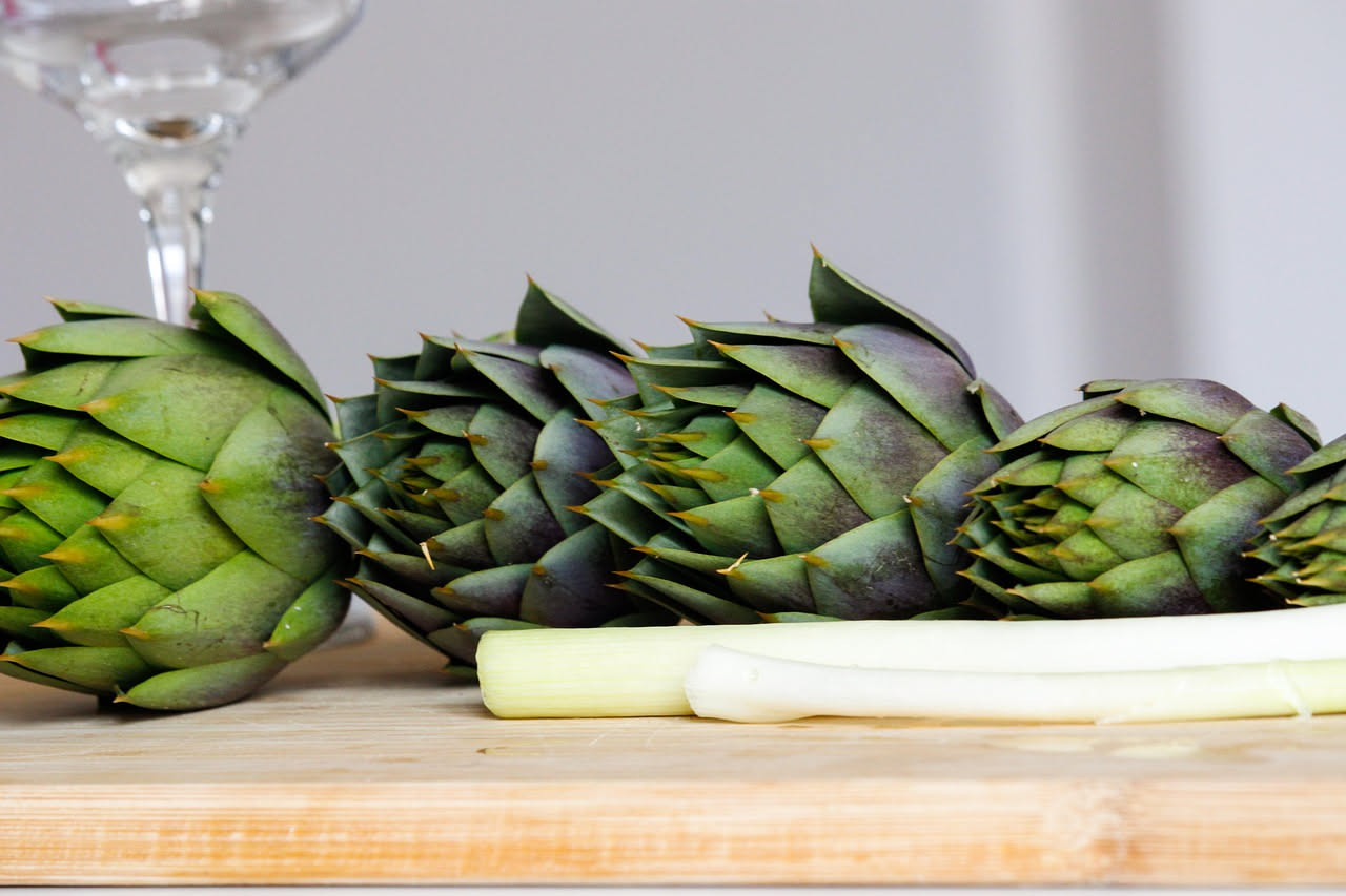Insalata di carciofi in ciotola bianca: fette sottili di carciofi crudi beige e verdi disposte su fondo scuro, con gocce di olio d'oliva lucido, scaglie di parmigiano bianche, qualche foglia di prezze